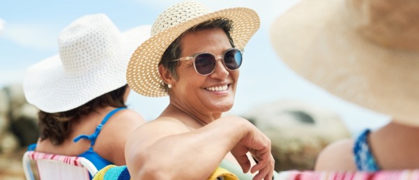 A woman sitting outside wearing a large sun hat and sunglasses smiles broadly