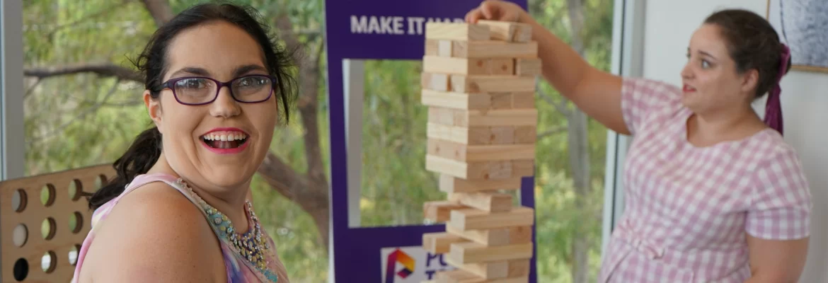Two women smile while playing massive jenga. A Power to You sign can be seen in the background