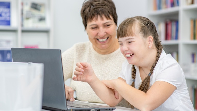 A young girl smiles at a laptop screen while an older woman sits next to her