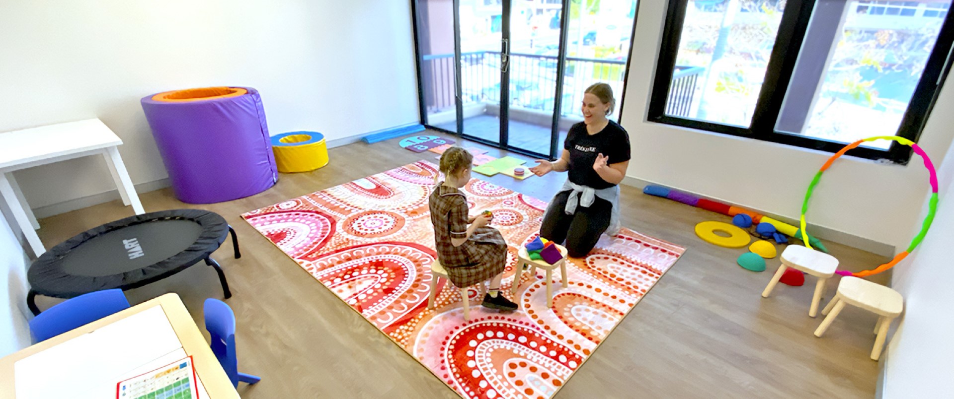A young woman and a young girl play on a large rug in an Allied Health therapy room