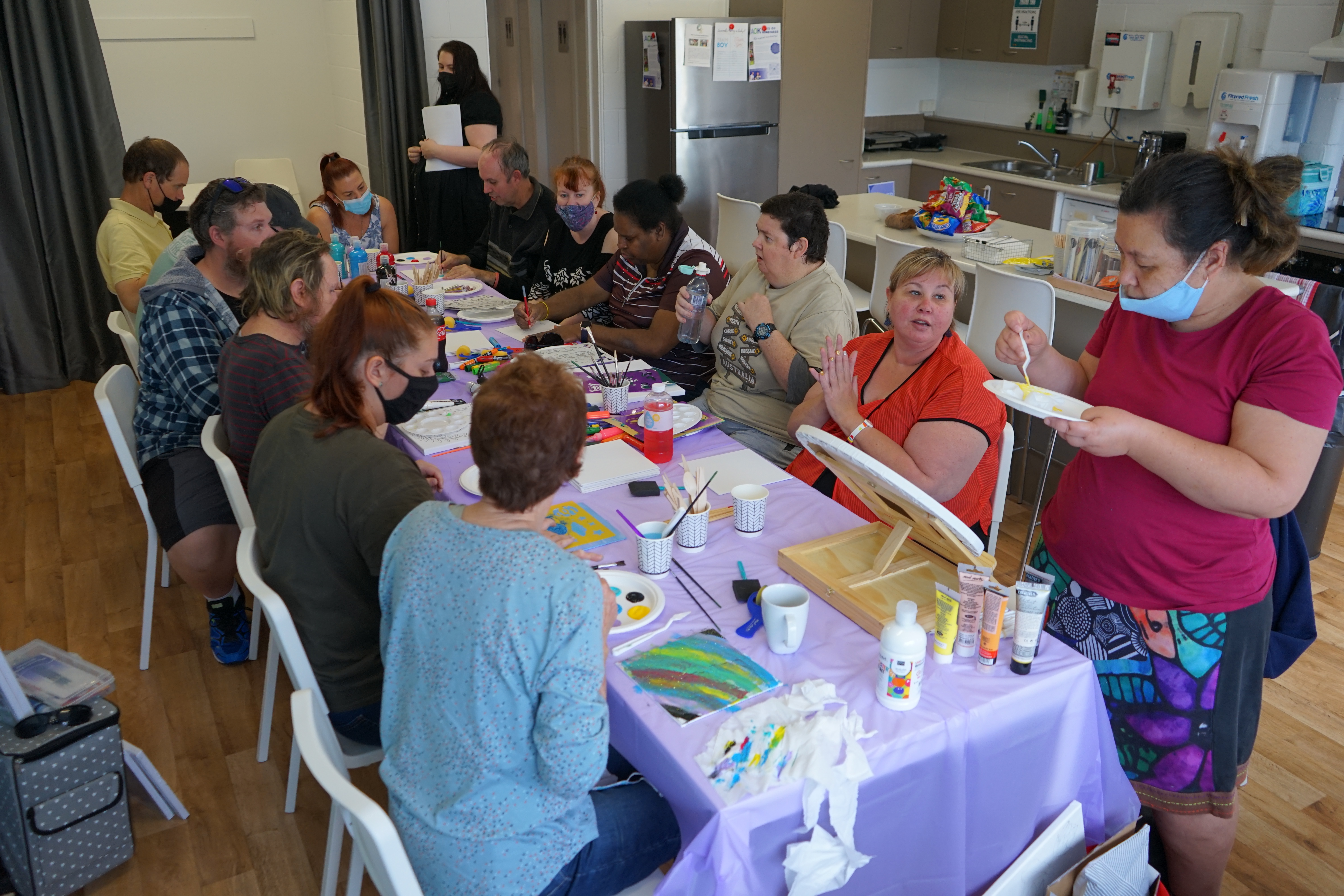 A group of people sit at a long table filled with painting equipment