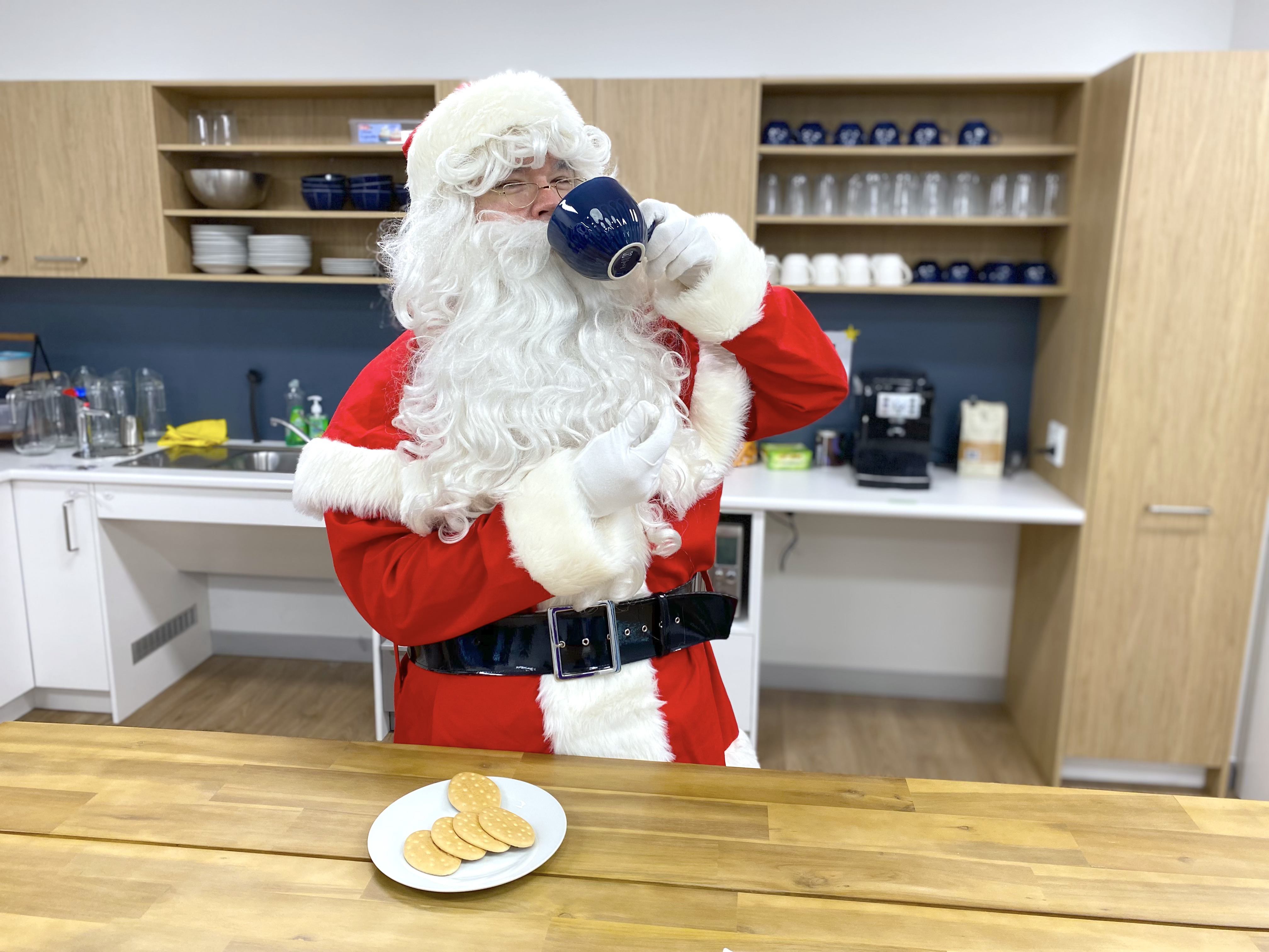 Santa in the Mamre kitchen drinking from a cup holding cookies