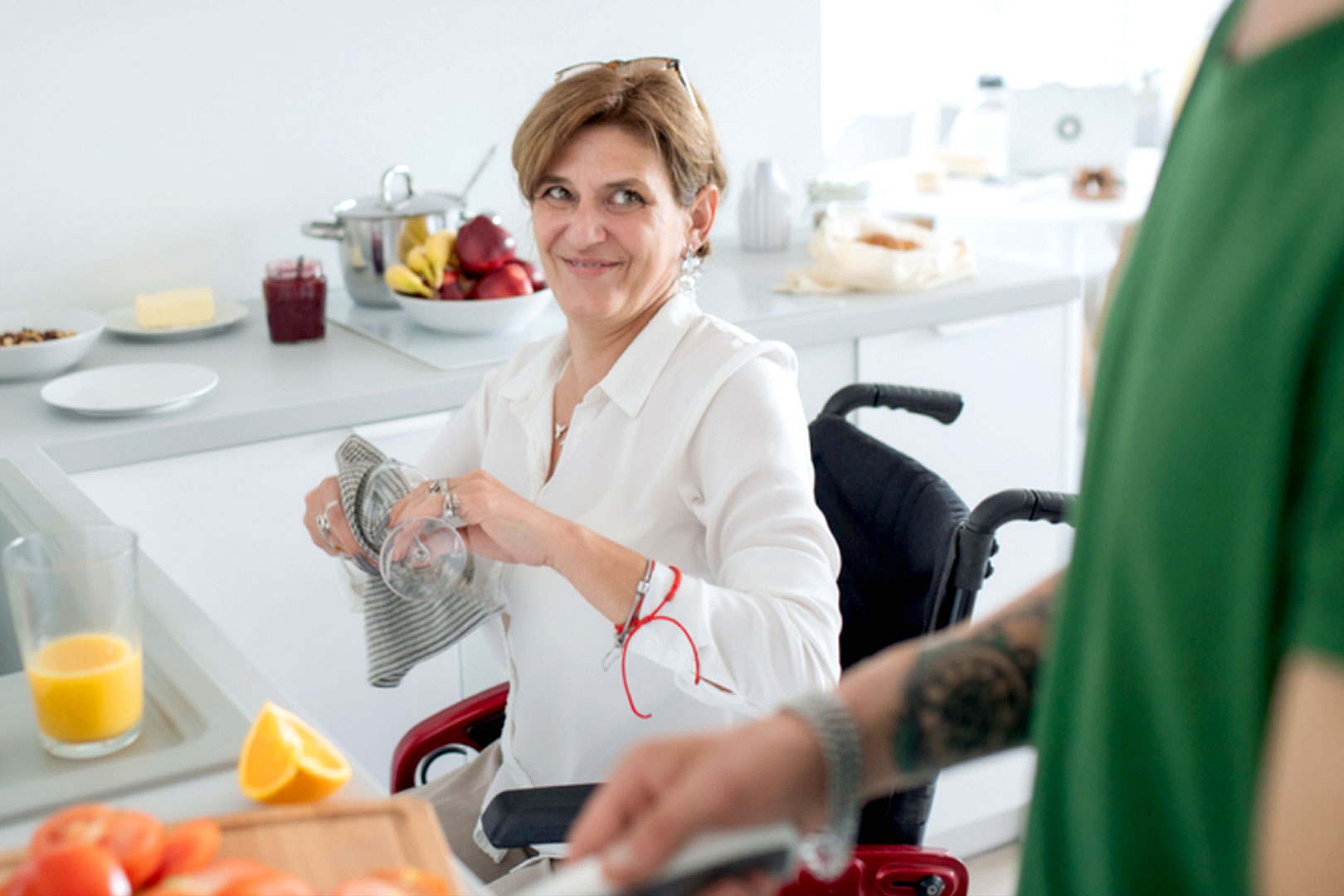 A woman in a wheelchair smiles at a man standing in a green top while she dries a cup with a dish towel in the kitchen