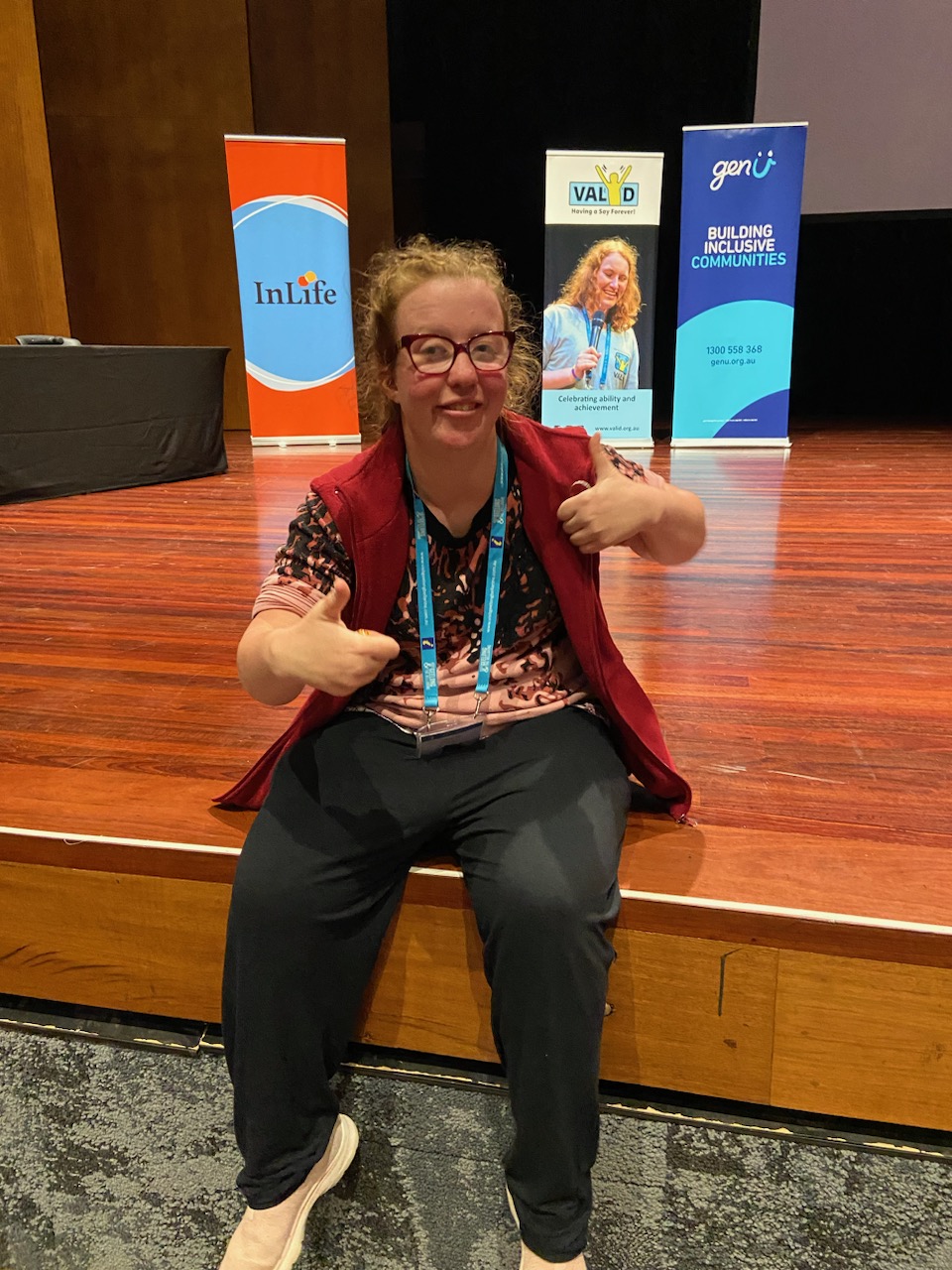 Jasmin sits on a stage at the VALID conference while smiling and holding two thumbs up
