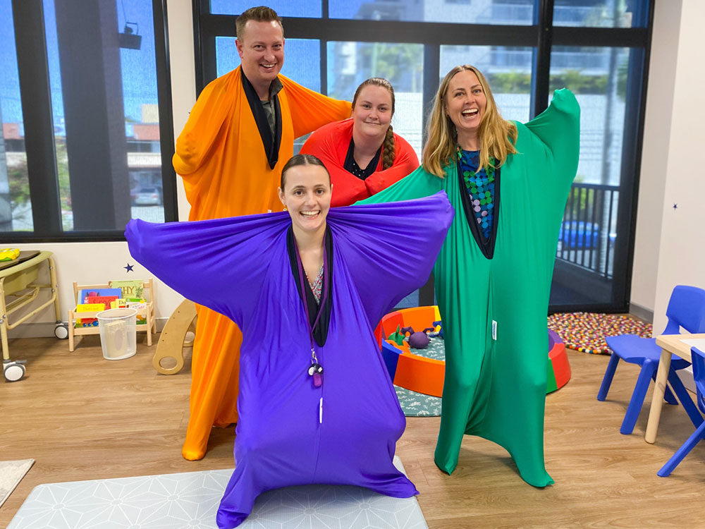 Four people standing in brightly coloured sensory body socks smiling. Mamre news logo in bottom right corner.
