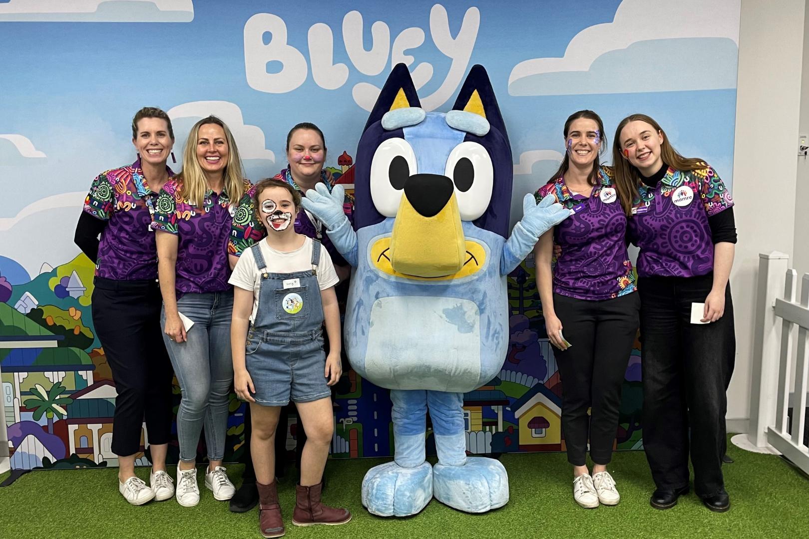 Mamre staff members and a young girl group around Bluey for a group photo