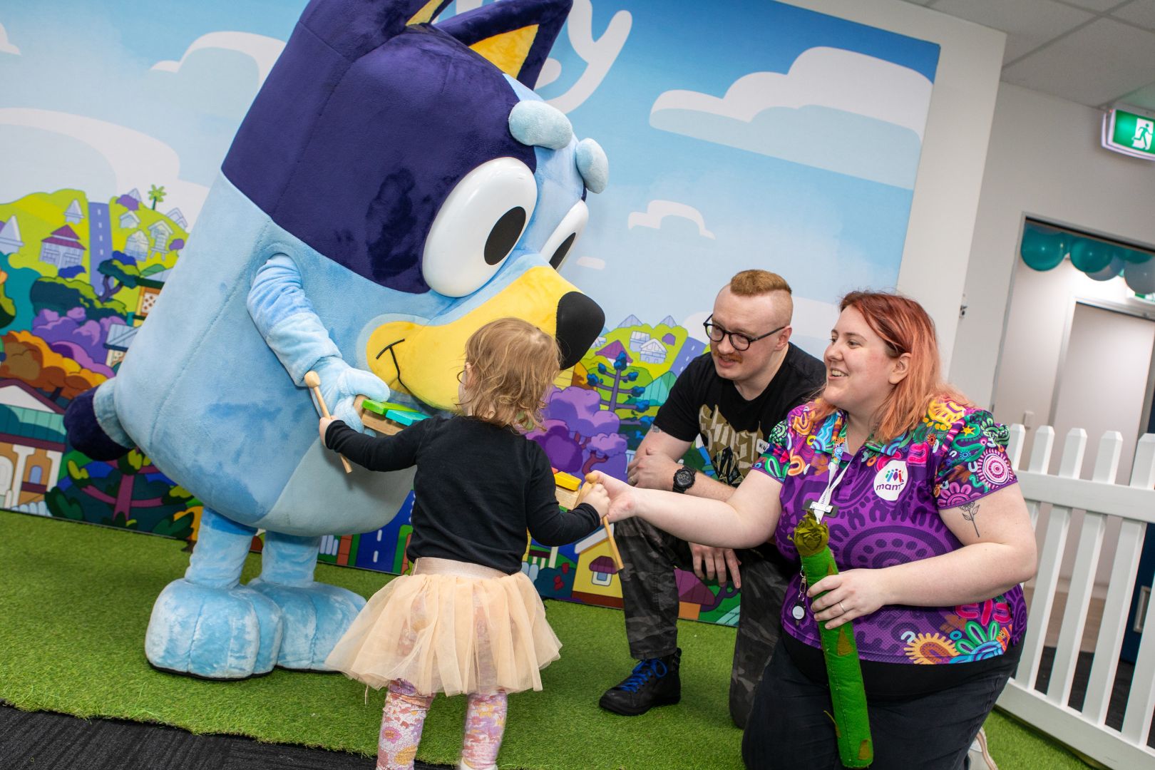 Bluey plays with a young customer while a man and a Mamre staff member watch on.
